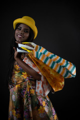 Fashion portrait of young dark skinned Indian Bengali brunette girl in floral dress and yellow cap holding bags in front of black copy space studio background. Indian fashion photography and lifestyle