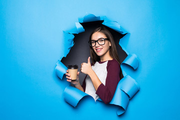 Young pretty woman holding coffee with thumbs up while looking through blue hole in paper wall.