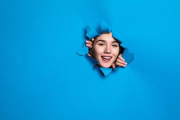 Young pretty woman face looking through blue hole in paper wall.