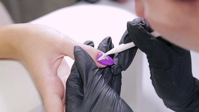 Manicurist woman in a beauty salon decorates client's nails with gel varnish with rhinestones.