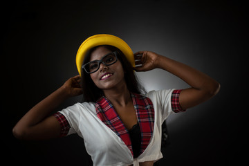 Portrait of an Asian/Indian/African brunette dark skinned young girl in sexy school uniform and spectacles with yellow hat in a black/grey studio background. Fashion and cosplay photography.