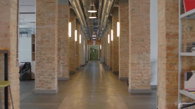 Zoom out shot of empty hallway with brick columns and lamps in modern loft office. There are comfortable bean bag chairs, shelves with stationery and chairs