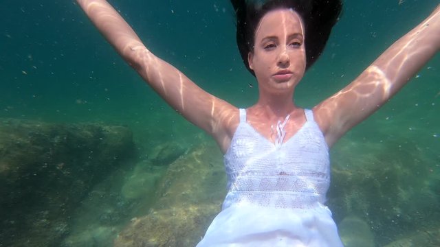 Beautiful young woman underwater in a white dress like an angel, artistic expression, dreamy, lifting her arms and opening her eyes, slow motion