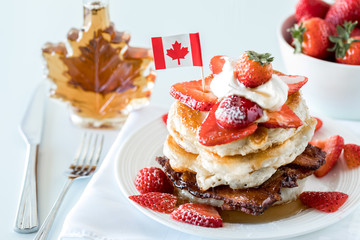 A close up of a stack of pancakes with a bottle of syrup and bowl of strawberries in behind. Canada Day concept.