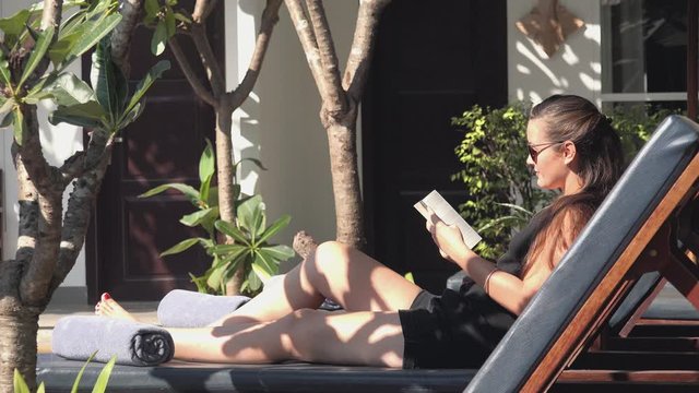 Medium Side Shot of a Young Woman Reading a Book by the Pool at a Tropical Hotel