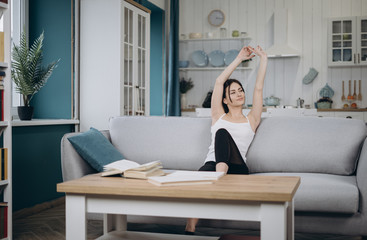 In cozy living room happy woman put hands behind head sitting leaned on couch 30s european female enjoy lazy weekend or vacation, housewife relaxing feels satisfied accomplish chores housework concept