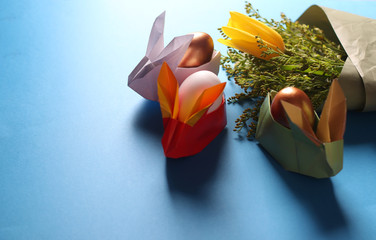Easter still life. Paper rabbits. Origami. Easter Golden eggs. On the blue table, a bouquet of...