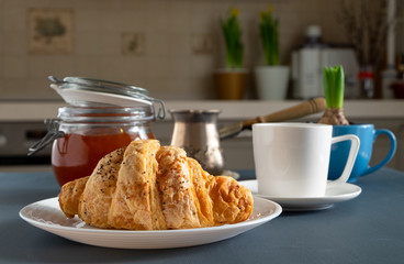Croissant, cup of coffee or tea, glass jar with jam, coffee maker and cultivated hyacinth bulb on the table in a kitchen. Breakfast, coffee break. Horizontal orientation. Selective focus. 