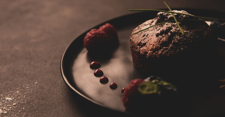 Czekoladowe babeczki z malinami i jagodami na czarnym talerzu z podświetleniem, Zdjęcie żywności z delikatnymi kolorami w stylu retro - 329643419