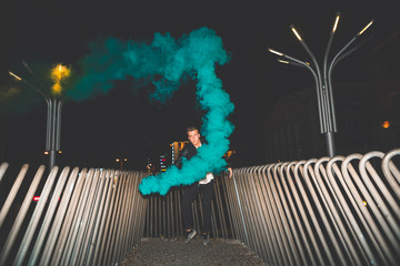 Black man in a leather jacket standing next to balcony. Colorful portrait of european teenager with blue smoke bomb. Night time.