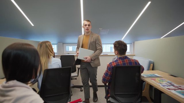 Tracking shot of young man with laptop walking down corridor, entering office and reporting bad news to business team listening to him and thinking of solution