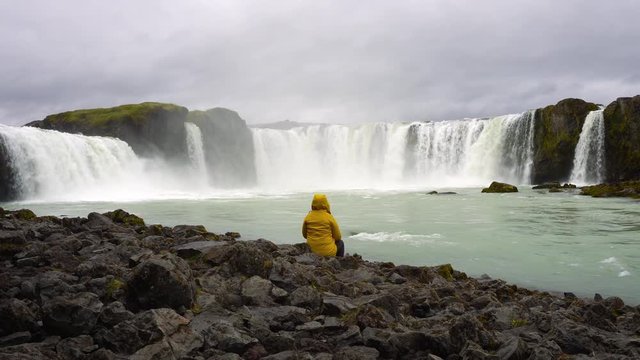Tourist in a yellow jacket relaxing at the Godafoss waterfall in Iceland