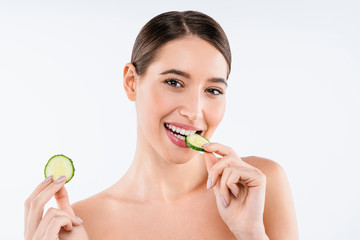 Beauty portrait of smiling young topless woman eating slice of cucumber standing isolated over white background