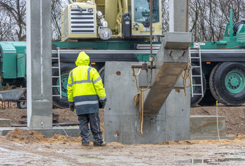 construction worker in a yellow raincoat supervises and controls the erection of a large concrete column
