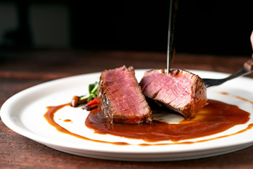 Beef tenderloin steak in demiglas sauce is cut with a knife on a white platter