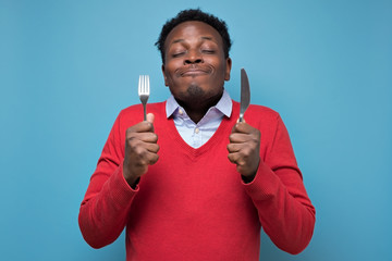 Young hungry crazy african american man in red sweater holding a fork and a knife. Isolated on blue...