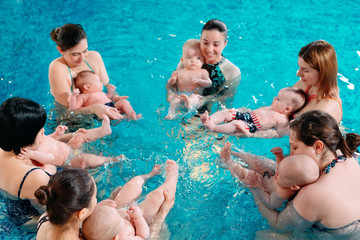 A group of mothers with their young children in a children's swimming class with a coach.