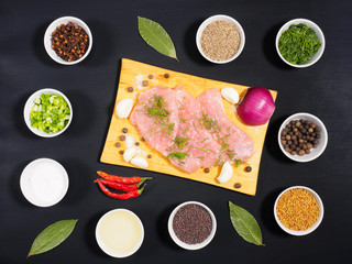 Sliced pieces of meat turkey game filet on a wooden cutting board, garlic, cumin (jeera), salt, fennel, allspice, fenugreek, mustard seeds, oil, green and red onion, cloves on black wooden background.