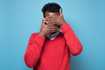 African american black man covering eyes being stressed trying to stay anonym. Studio shot on blue wall.