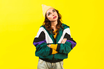 Modern beautiful girl in oversized sports jacket, hat and jeans, Billie Eilish style, back in the 90s, 2000s. Posing in the studio on a yellow background, strong funny emotions