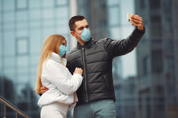 Fototapeta premium Boyfriend and girlfriend are making selfie and wearing disposable masks. They are standing on the street near a skyscraper.
