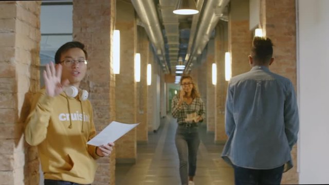Multiethnic colleagues walking through hallway, smiling and saying hi at camera. Caucasian lady going with coffee, Asian guy reading paper, mixed raced woman talking on phone and man riding scooter