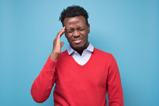 Serious afro american man in red sweater with headache in studio on blue isolated background