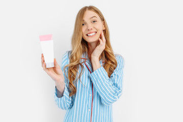 Portrait of happy beautiful young woman in pajamas standing isolated on white background applying face lotion