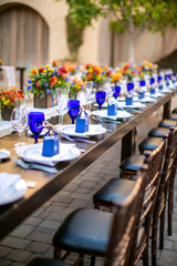 Table setting for a wedding celebration in an outdoor mission style patio with blue and orange theme, and flower decorations