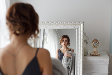 Beautiful red-haired woman in a black elegant dress looks in the mirror