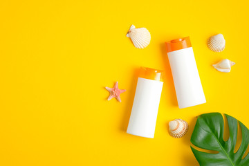 Two white plastic sunscreen lotion bottles, seashells and tropical leaf on yellow background. Sun...