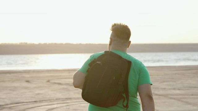 A plump guy in a green T-shirt goes to the water, throwing a backpack over his shoulder.