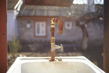 Old rusty faucet on a sink in a rural yard. The facade of the house in the background in blur.