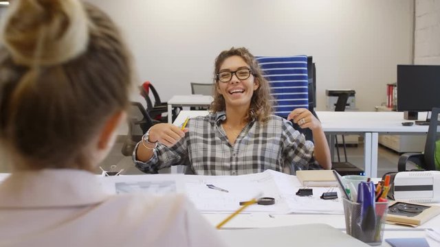 Young cheerful mixed-raced woman in casual outfit sitting with drawing tools and paper at desk, smiling and discussing interior designs with female coworker while cooperating in open space office