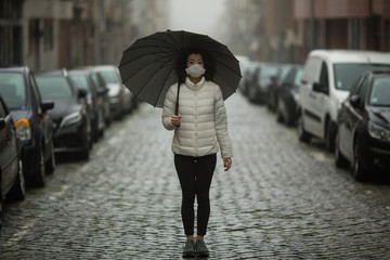 Pandemic. A multicultural asian woman in antiviral mask stands in the street in cloudy weather.