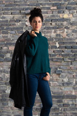 Naklejka premium Portrait of young attractive woman in sweater with leather jacket on her shoulder. Brunette female model with topknot hairstyle looking at camera. Old brick wall background...