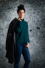 Young attractive brunette woman portrait. Female model with topknot hairstyle wearing sweater and holding leather jacket on her shoulder. looking at camera. Old dirty wall background...