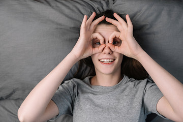 Cheerful funny young woman having fun making glasses shape with hands, lying in comfortable bed, looking at camera, smiling female wearing grey t-shirt doing funny face. People emotions concept
