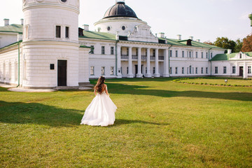 The bride in a white wedding dress walks on a green lawn against the background of a white historical building, a beautiful sunset, author's wedding dress, the back view of the bride, chic black curls