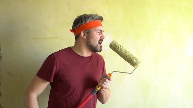 Bearded man against the wall. Sings in a construction roller. Performance of the artist. Musical number. On the head is an orange ribbon. Repair work in the room. Frick. Retro style. Green wall. Rock-