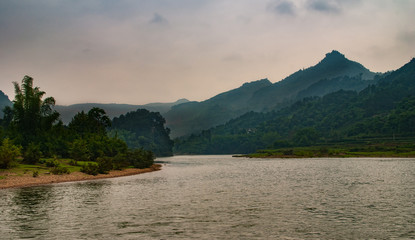 Guilin, China - May 10, 2010: Along Li River. Wide landscape over light reflecting water with tall karst mountains in back and green trees on flat brown rocky shore.