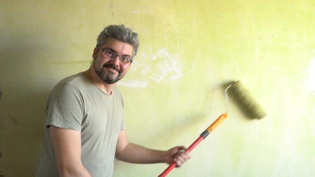 A bearded man paints a wall. Good mood. A smile on his face. Repair work in the room. Actor. Frick. Clown.