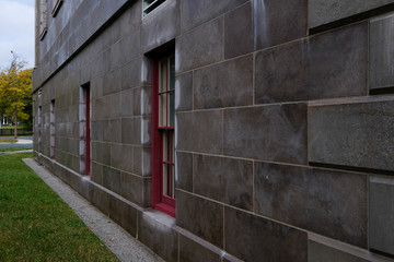 A large block wall of a vintage building with double hung windows on the ground level. The bricks are a dark grey.There's grass on the ground and trees in the background.The wooden windows are closed.