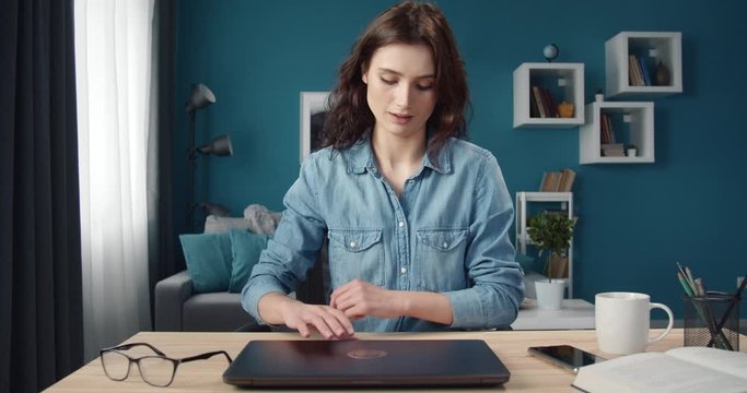 Cheerful young lady in casual clothing opening personal computer to start working at own apartment. Beautiful lady with curly hair sitting at table with laptop.