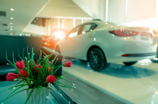 Fake red flowers in vase decorated on table in reception area in showroom. Artificial flower on blurred background of new and shiny white luxury car parked in modern showroom for sale. Car dealership.