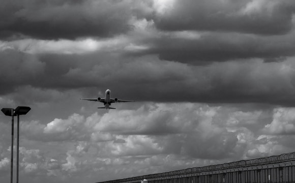 Black and white scene of airplane landing at the airport. Commercial airline flying on dark sky and white clouds over fence and lamp post. Aviation business crisis concept. Journey vacation flight.
