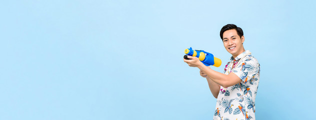 Smiling handsome Asian man playing with water gun isolated on banner blue background with copy...