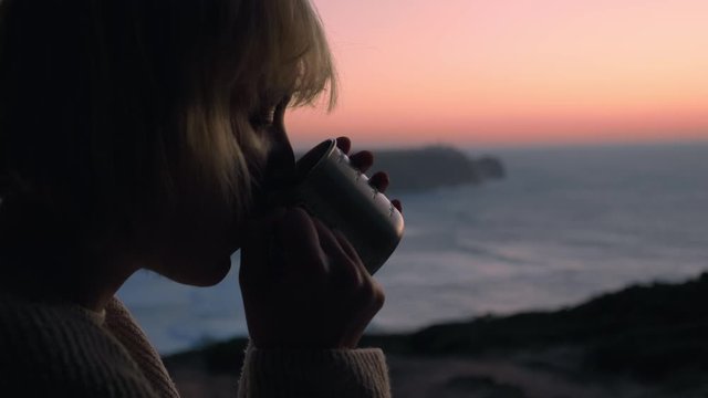 Close up shot of a female face with blonde messy hair sitting and relaxing in her Camper van drinking hot tea / coffee. A van is parked on a cliff with a beautiful view on the sunset/sunrise. 