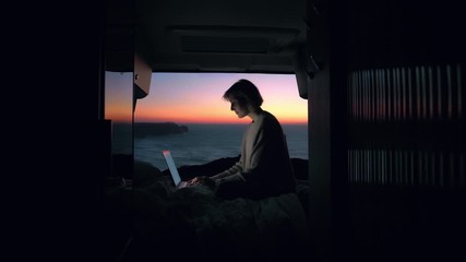 A young entrepreneur woman is working on her computer in a camper van. The door is open to an epic view of the sea in a sunset / sunrise. Remote working anywhere, the concept of digital nomad. - Powered by Adobe