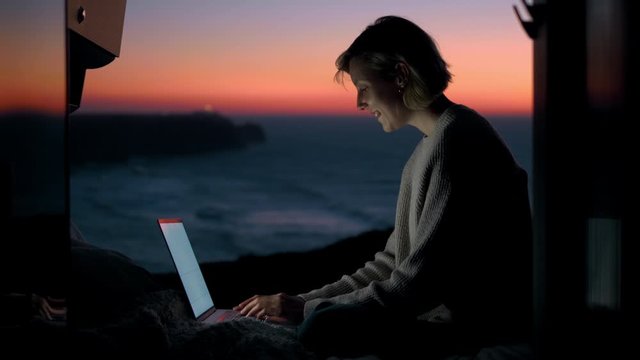 A young entrepreneur woman is working on her computer in a camper van. The door is open to an epic view of the sea in a sunset / sunrise. The lighthouse is in the background. Remote working anywhere.